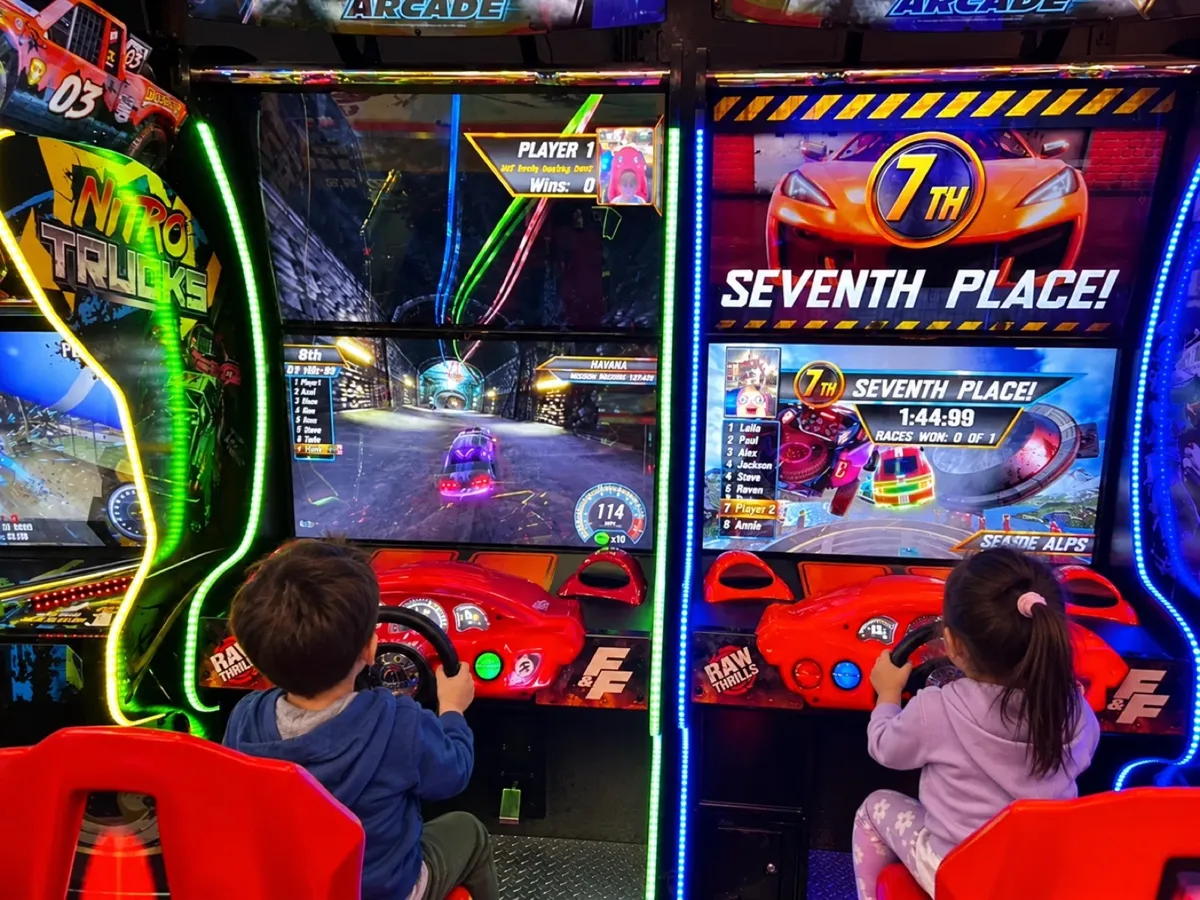 Two kids seen from behind racing each other on full-size arcade driving games in the rental's game room