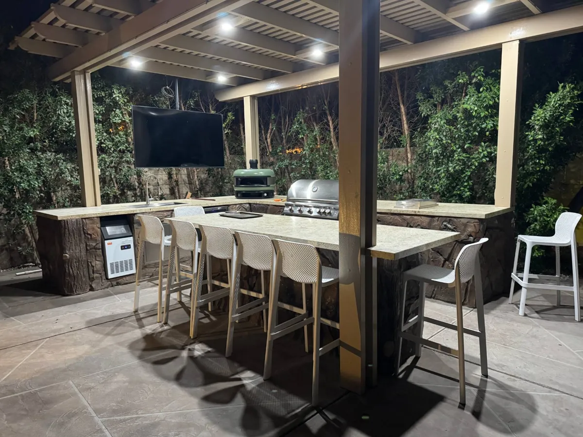 Covered outdoor kitchen with a grill, pizza oven, sink, ice maker, TV, and white bar stools around a stone counter